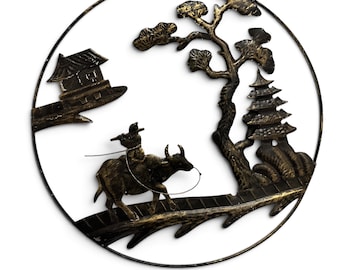 May include: A circular black and gold metal wall hanging featuring a silhouette scene of a person riding a water buffalo, a small house on a ledge, a tall pagoda, and a tree, all enclosed within a thin metal ring frame.