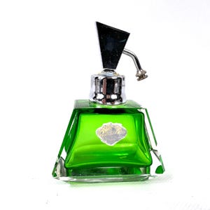 May include: A vintage green glass perfume bottle with a chrome-colored geometric top and sprayer. The bottle has a square base and a label with text. The perfume bottle is on a white background.