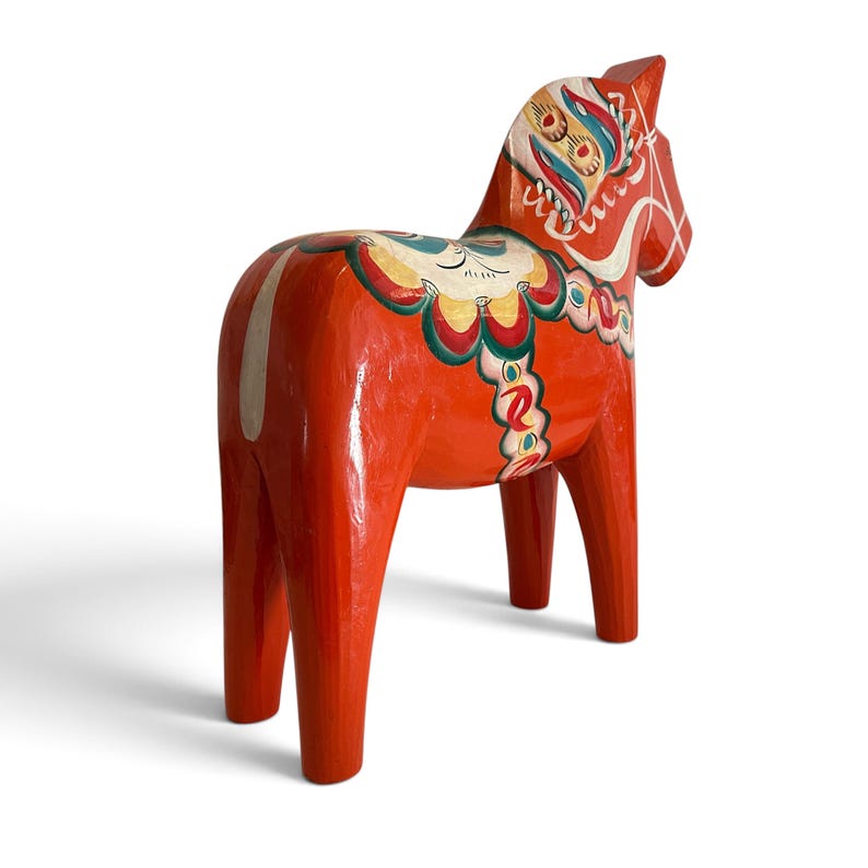 Puede incluir: Una figura de caballo de madera de color naranja brillante con patrones decorativos pintados a mano en blanco, amarillo, verde y azul. El caballo tiene un acabado liso y brillante con motivos florales complejos en el lomo y el cuello.