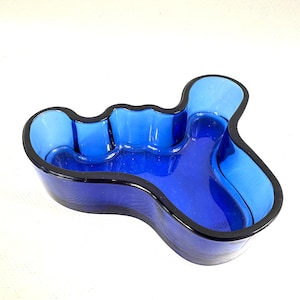 May include: A cobalt blue glass bowl with an irregular, organic, free-form shape. The bowl features a thick, dark blue rim that follows the undulating, wavy contours of the vessel, creating a deep, open basin with smooth, curved interior walls.