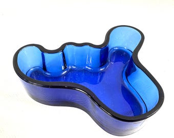May include: A cobalt blue glass bowl with an irregular, organic, free-form shape. The bowl features a thick, dark blue rim that follows the undulating, wavy contours of the vessel, creating a deep, open basin with smooth, curved interior walls.