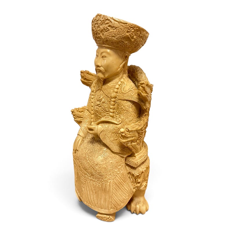 Op de afbeelding: Een cr&egrave;mekleurig uitgesneden beeldje van een zittende figuur met een versierde hoed en traditionele gewaden met complexe drakenmotieven. Het beeldje staat op een voetstuk met drakenkoppen en klauwen, met gedetailleerd reli&euml;fwerk.