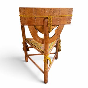 May include: A wooden corner chair featuring intricate carvings on the backrest and frame. The chair includes a triangular seat cushion with a patterned fabric and is accented with thick, braided mustard yellow rope ties and tassels.