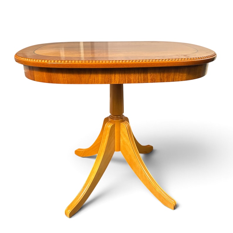 Op de afbeelding: Een ovale houten tafel op een voetstuk met een gepolijst bruin oppervlak en een decoratieve kralenrand. De tafel heeft een centrale kolomsteun die uitloopt in drie gebogen poten, afgewerkt in een warme, lichtgekleurde houtnerf.