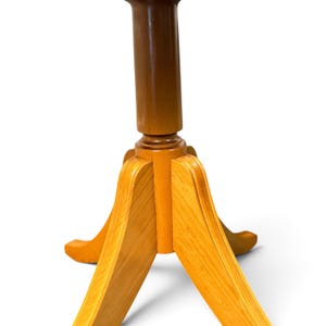Op de afbeelding: Een houten voetstuk voor een tafel of standaard, bestaande uit een centrale cilindrische kolom ondersteund door drie gebogen, uitlopende poten. Het hout heeft een gladde, licht- tot middenbruine afwerking met zichtbare houtnerven.