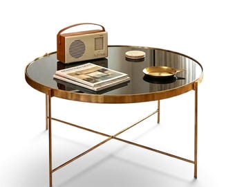 May include: A round coffee table with a dark glass top and a thin gold metal frame featuring a cross-base design. On the table sits a beige radio with a brown handle, a stack of magazines, a small round coaster, and a shallow gold metal dish.