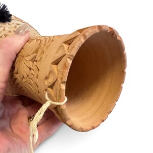 May include: A light brown, hollow, cylindrical clay drum with a flared base and a wide, open top. The exterior surface features intricate, hand-carved floral and geometric patterns. A thin, light-coloured string is attached to the side.