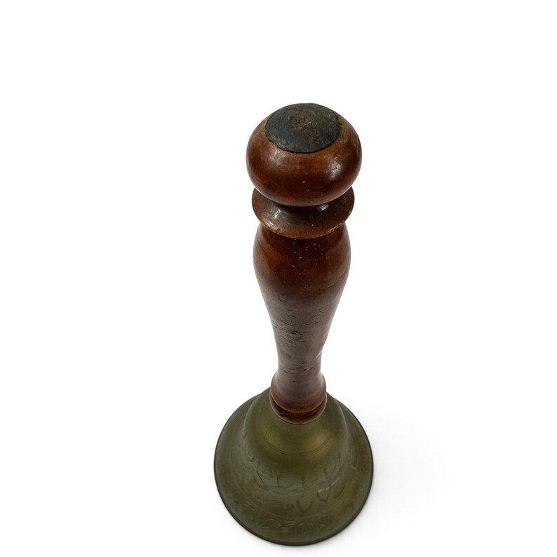 May include: A hand bell featuring a dark brown wooden handle with a rounded top knob and a flared brass base. The brass bell section displays a subtle, engraved floral pattern around its circumference, showing a weathered, aged metal finish.