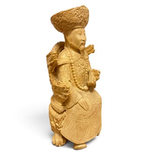 Op de afbeelding: Een cr&egrave;mekleurig uitgesneden beeldje van een zittende figuur met een versierde hoed en traditionele gewaden met ingewikkelde drakenmotieven. De figuur heeft een lange baard, draagt een kralenketting en zit op een voetstuk met gebeeldhouwde drakenkoppen.