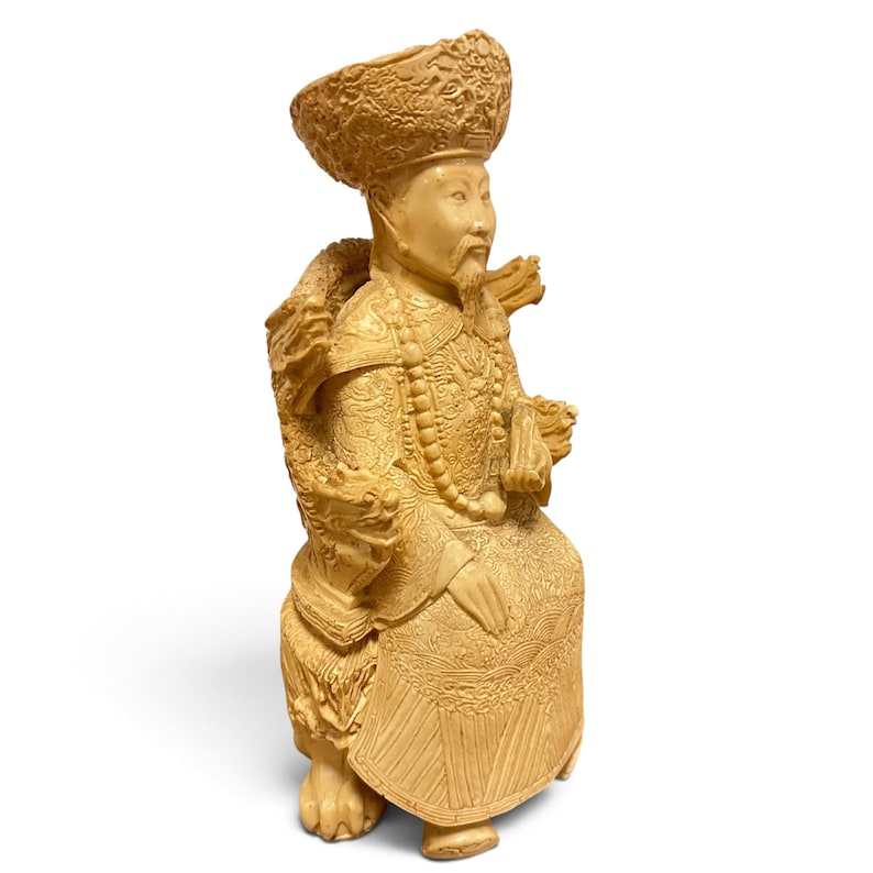 Op de afbeelding: Een cr&egrave;mekleurig uitgesneden beeldje van een zittende figuur met een versierde hoed en traditionele gewaden met ingewikkelde drakenmotieven. De figuur heeft een lange baard, draagt een kralenketting en zit op een voetstuk met gebeeldhouwde drakenkoppen.