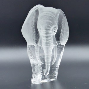 May include: A clear glass elephant sculpture with detailed trunk and tusks. The sculpture has textured lines, giving it a frosted appearance. The elephant is standing upright, with its ears flared out. The sculpture is on a dark background.