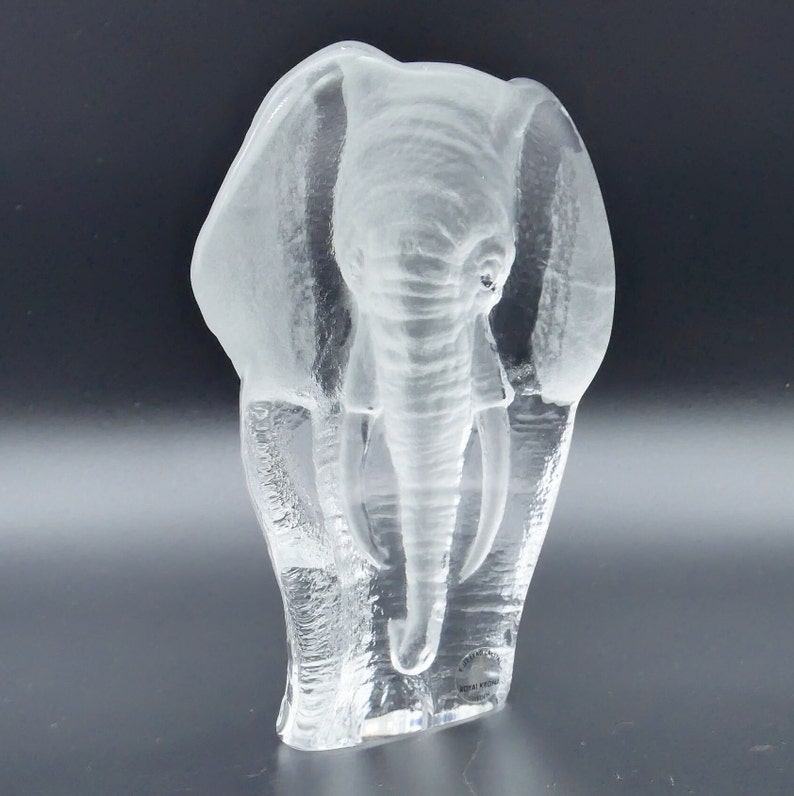 May include: A clear glass elephant sculpture with detailed trunk and tusks. The sculpture has textured lines, giving it a frosted appearance. The elephant is standing upright, with its ears flared out. The sculpture is on a dark background.