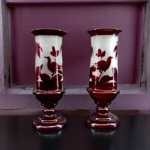 May include: Two matching glass vases with a white interior and a dark red exterior. The vases feature a bird and floral design. The vases have a flared rim and a pedestal base. The vases are displayed on a dark surface.