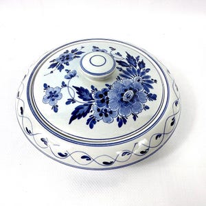 May include: A round, white ceramic serving dish with a lid, decorated with blue floral patterns. The lid has a small knob and a matching floral design. The rim of the dish is decorated with a delicate blue and white pattern.