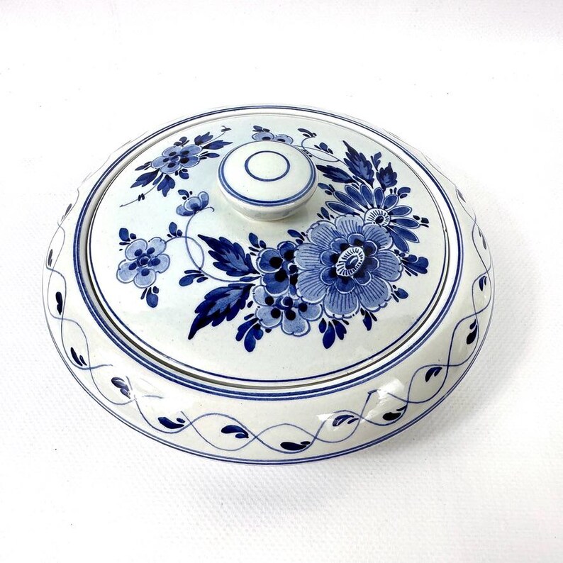 May include: A round, white ceramic serving dish with a lid, decorated with blue floral patterns. The lid has a small knob and a matching floral design. The rim of the dish is decorated with a delicate blue and white pattern.