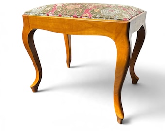 May include: A wooden vanity stool with four curved legs and a rectangular seat upholstered in a fabric featuring a floral pattern in shades of red, green, and beige. The wooden frame has a smooth, polished light brown finish.