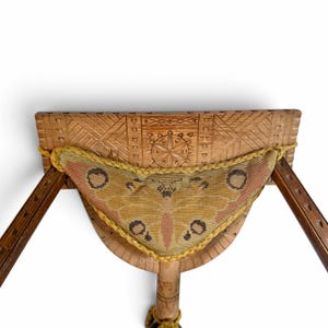 May include: A wooden stool featuring a carved geometric pattern on the seat and a triangular needlepoint cushion attached with yellow braided trim. The cushion displays a moth-like pattern in shades of olive green, brown, and muted orange.