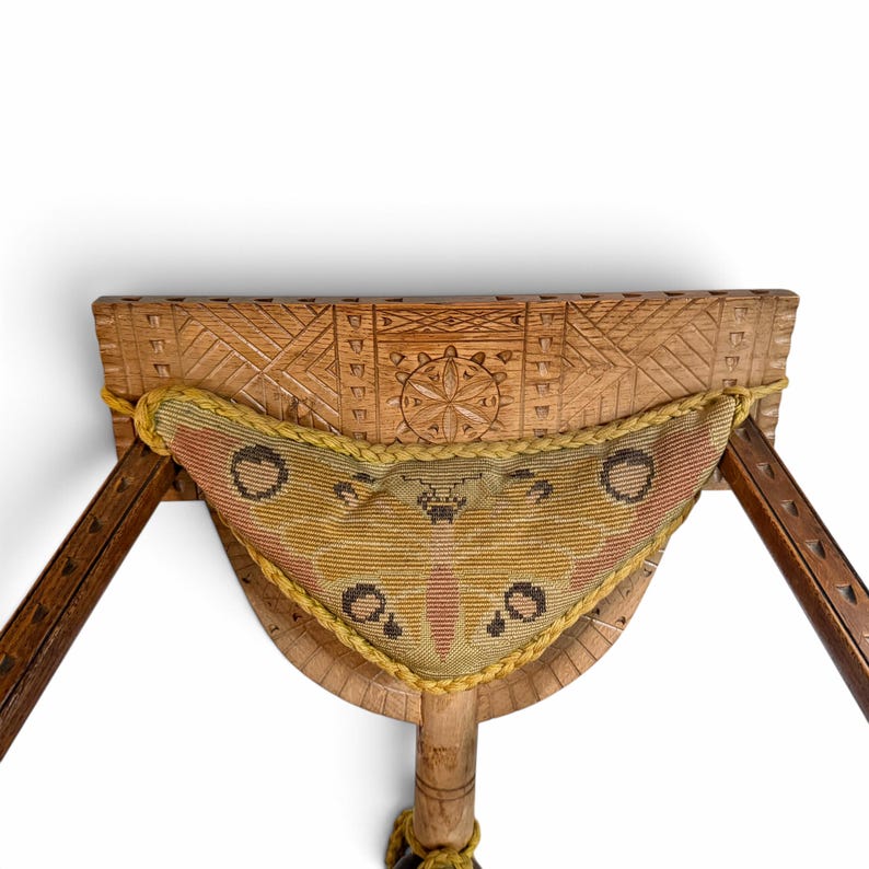May include: A wooden stool featuring a carved geometric pattern on the seat and a triangular needlepoint cushion attached with yellow braided trim. The cushion displays a moth-like pattern in shades of olive green, brown, and muted orange.