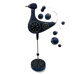 May include: A dark blue wooden bird figurine standing on a thin metal rod and a circular base. The bird features painted green and white circular patterns on its body and base, with a tail made of thin wires ending in small dark blue spheres.