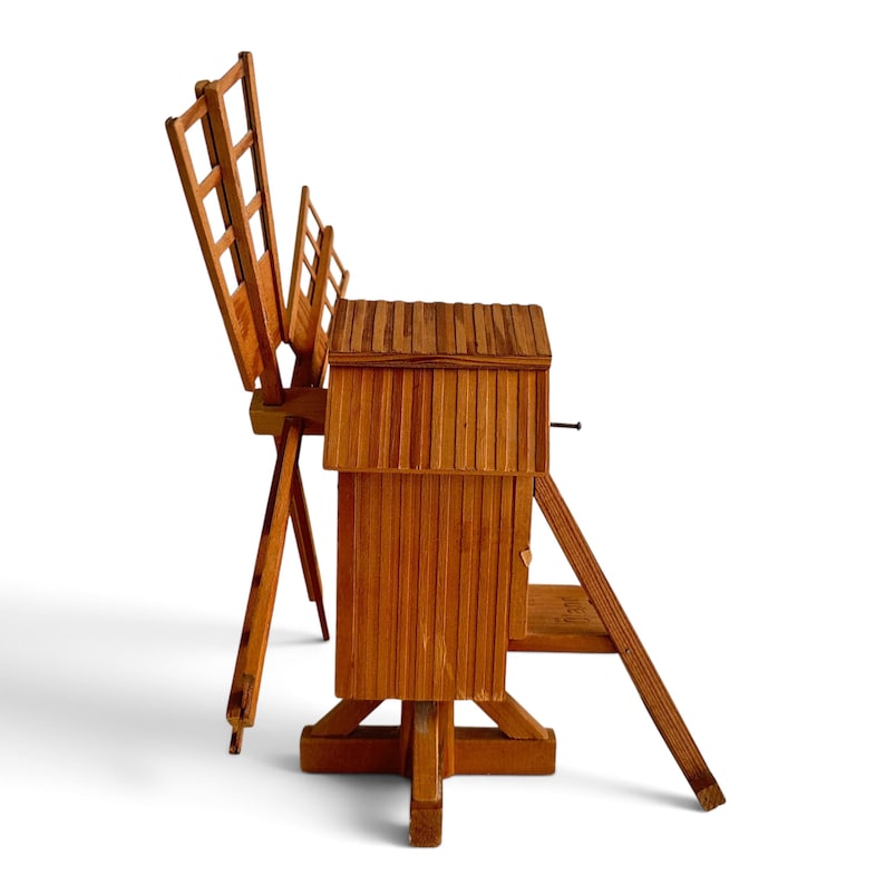 May include: A wooden model of a windmill featuring a rectangular main structure with vertical slats, a sloped roof, and a rotating blade assembly attached to a support frame. The object is constructed from light brown wood with visible grain.