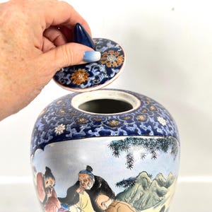 Puede incluir: Un tarro de jengibre de cer&aacute;mica con una escena pintada a mano de figuras en un paisaje con monta&ntilde;as y bamb&uacute;. El tarro tiene una base blanca con detalles en azul, naranja y verde. Se sostiene una tapa a juego con un remate azul sobre el tarro.