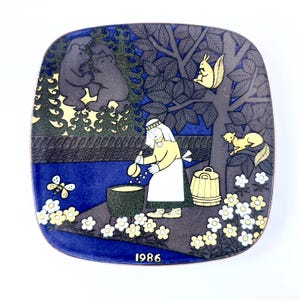 Op de afbeelding: Vierkante decoratieve plaat met een grillige bossc&egrave;ne. De plaat toont een vrouw die in een pot roert, beren, eekhoorns en een vlinder. Het kleurenpalet omvat blauw, geel en grijs. Het jaar "1986" staat onderaan gedrukt.