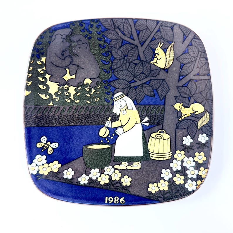 Op de afbeelding: Vierkante decoratieve plaat met een grillige bossc&egrave;ne. De plaat toont een vrouw die in een pot roert, beren, eekhoorns en een vlinder. Het kleurenpalet omvat blauw, geel en grijs. Het jaar "1986" staat onderaan gedrukt.
