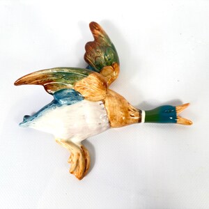 May include: A ceramic mallard duck wall plaque in a flying position with wings extended. The duck features a glossy finish with a dark green head, a white neck ring, tan and brown body feathers, and iridescent blue and green wing accents.