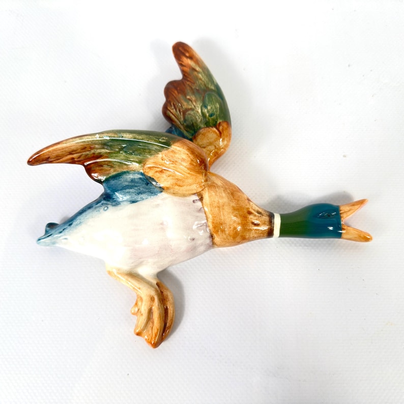 May include: A ceramic mallard duck wall plaque in a flying position with wings extended. The duck features a glossy finish with a dark green head, a white neck ring, tan and brown body feathers, and iridescent blue and green wing accents.