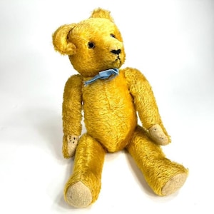 May include: A vintage golden yellow teddy bear with a blue ribbon around its neck. The bear has black eyes and tan-colored paws and feet. The plush toy is sitting with its legs extended and arms at its sides.
