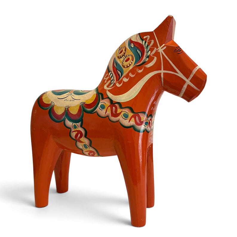 Puede incluir: Una figura de caballo de madera tallada pintada en un tono naranja vibrante. El caballo presenta intrincados patrones florales y geom&eacute;tricos pintados a mano en tonos azul, amarillo, verde y blanco en la crin, el lomo y los costados.