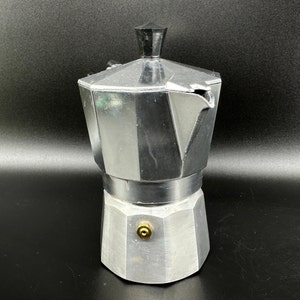 Puede incluir: Una cafetera de metal plateado con un mango negro y un pico. La cafetera tiene un cuerpo redondeado y una base plana.