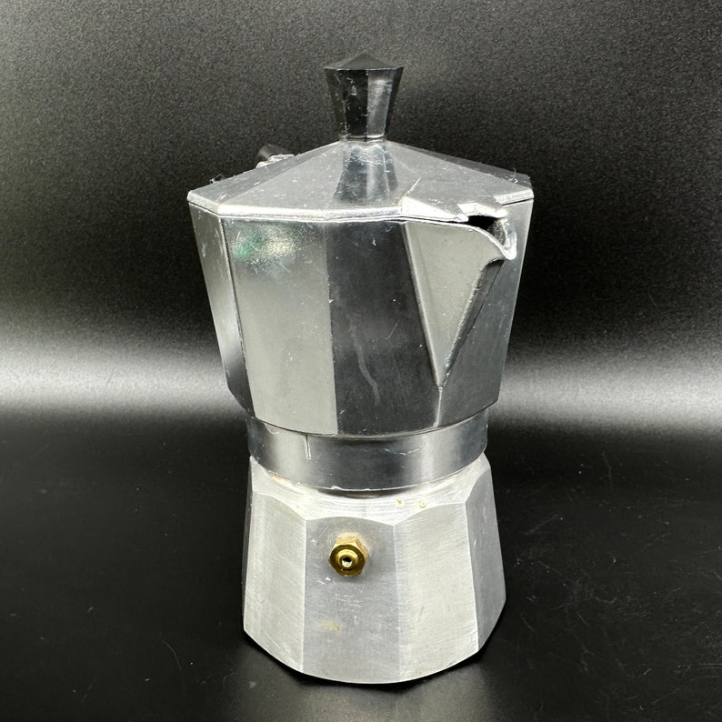 Puede incluir: Una cafetera de metal plateado con un mango negro y un pico. La cafetera tiene un cuerpo redondeado y una base plana.