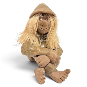 May include: A ceramic figurine of a seated figure with a smooth, light brown finish. The figure wears a pointed hat and has long, straight, light-colored fibrous hair. It is depicted in a sitting position with knees pulled toward the chest.