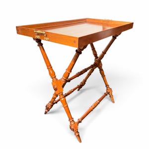 Puede incluir: Una mesa bandeja rectangular de madera con un acabado liso y pulido y un borde elevado. La bandeja cuenta con asas de metal dorado ornamentadas en los laterales y se apoya sobre una base plegable en cruz con patas torneadas.