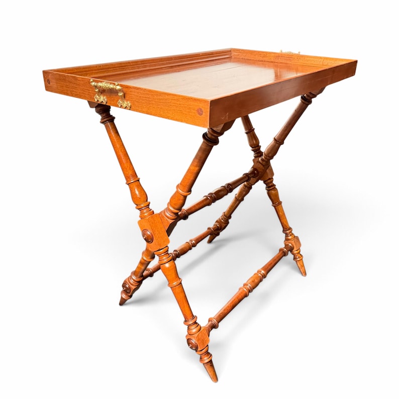 Puede incluir: Una mesa bandeja rectangular de madera con un acabado liso y pulido y un borde elevado. La bandeja cuenta con asas de metal dorado ornamentadas en los laterales y se apoya sobre una base plegable en cruz con patas torneadas.