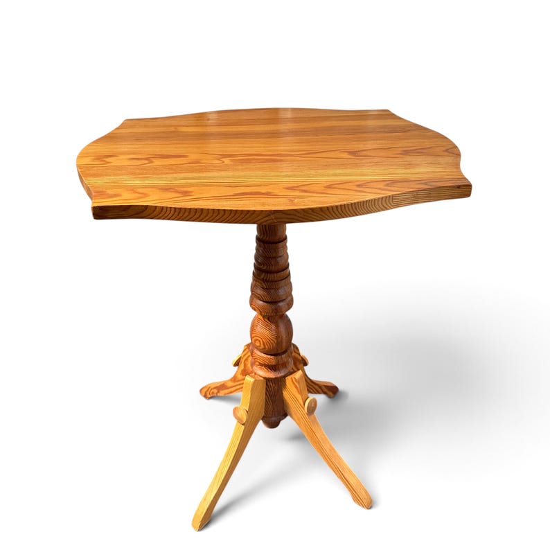 May include: A wooden side table featuring a light brown grain pattern with a smooth, irregularly shaped tabletop. The table is supported by a central, vertically turned pedestal column that rests on a three-legged base with curved feet.