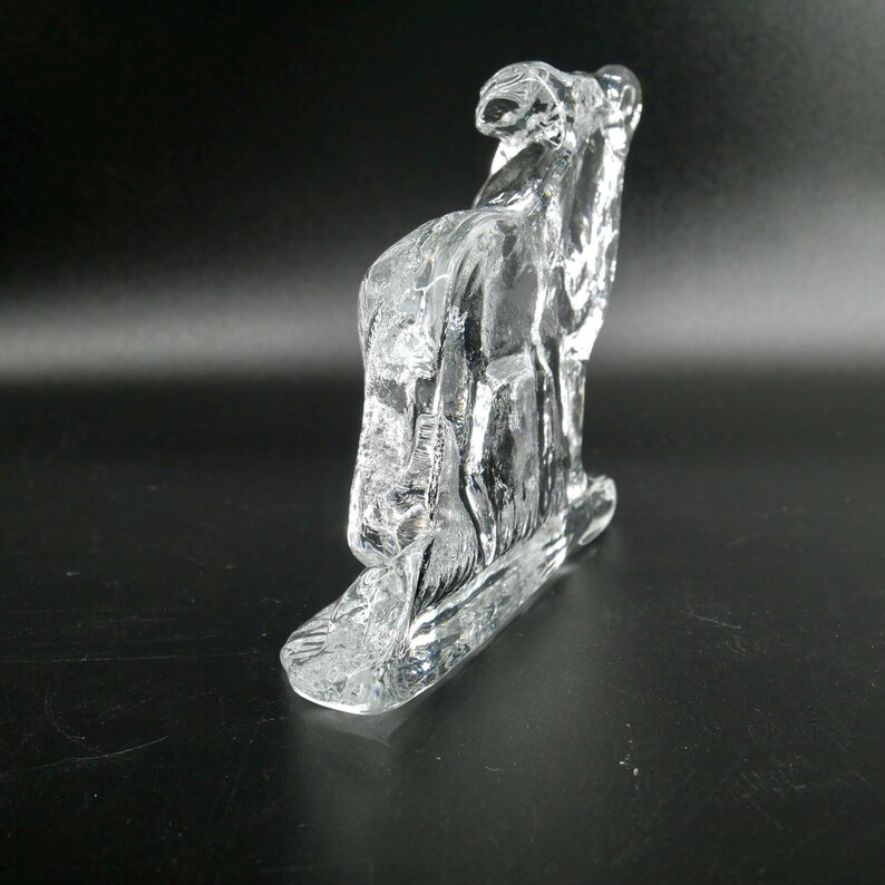 May include: A clear glass sculpture of a camel, with a textured surface. The sculpture is standing on a rectangular base. The camel is facing to the left.