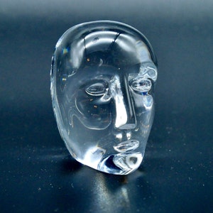 May include: A clear glass sculpture of a human face. The face has closed eyes, a prominent nose, and a defined chin. The sculpture is smooth and reflective, with light refracting through the glass.