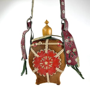 May include: A brown leather flask with a wooden stopper and a decorative red floral design. The flask has a braided leather trim and a burgundy leather strap with floral cutouts. The flask measures approximately 20 cm in height.