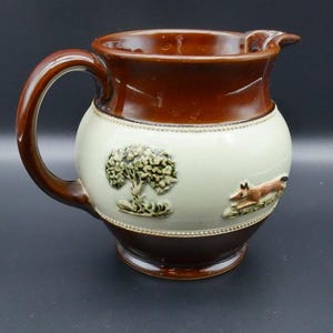 Può includere: Un'anfora in ceramica marrone e bianca con un design di albero e volpe. L'anfora ha un manico marrone e un bordo marrone.