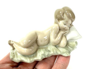 May include: A glossy porcelain figurine of a reclining figure resting their head on a white pillow. The figure has a smooth, light beige finish and is positioned on a textured, pale green base that resembles grass, with a reflective glazed surface.