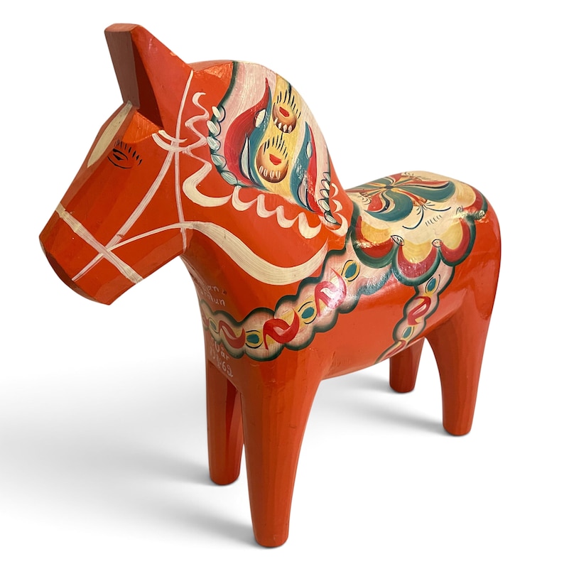 Puede incluir: Una figura de caballo de madera de color naranja brillante con patrones florales y geom&eacute;tricos pintados a mano en blanco, azul, verde y amarillo. El caballo tiene una crin estilizada, un dise&ntilde;o de silla de montar en el lomo y bridas pintadas, con texto visible.