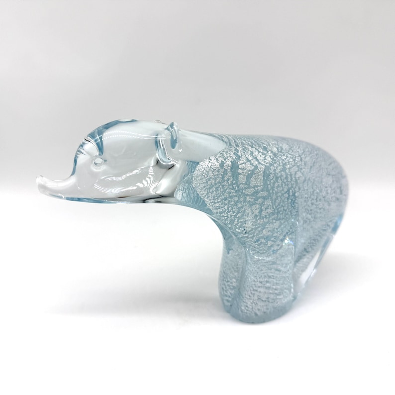 May include: A glass figurine shaped like a bear in a walking pose. The head is made of clear, smooth glass, while the body features a textured, light blue and silver metallic finish that creates a shimmering, crackled appearance.