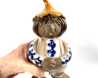 May include: A ceramic figurine of a gnome with a brown matte finish, wearing a tan pointed hat and a white shirt featuring blue floral patterns. The figure has dark brown hair, a round nose, and a single visible tooth, standing on two feet.
