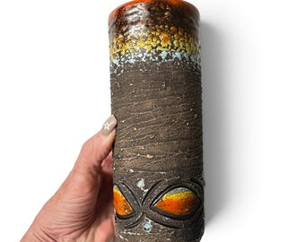 May include: A cylindrical ceramic vase with a dark brown, textured, bark-like surface. The top rim features a vibrant drip glaze in orange, yellow, and light blue. The lower section displays two carved, eye-shaped motifs filled with orange glaze.