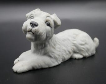 May include: A white ceramic figurine of a dog in a lying position. The dog has textured fur, pointed ears, and a dark gray nose and eyes. The sculpture is crafted with detailed facial features, including whiskers and eyebrows, and is positioned facing upward.