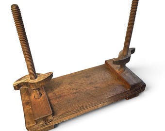 May include: A vintage wooden book press featuring a rectangular base and two vertical threaded metal rods. Each rod is equipped with a manually adjustable wooden handle, allowing for the compression of items placed between the two wooden plates.