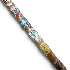 Puede incluir: Un bast&oacute;n de madera de color marr&oacute;n claro adornado con una serie de escudos de recuerdo de metal. Cada escudo presenta ilustraciones coloridas de paisajes monta&ntilde;osos, lagos y monumentos con textos que incluyen Triberger, Via Mala y Hochtannberg Pass.