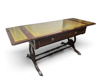 Puede incluir: Una mesa rectangular de madera oscura con dos alas abatibles y una superficie de cuero verde con bordes dorados en relieve. La mesa tiene dos cajones con tiradores metálicos redondos y se apoya en una base en forma de lira con patas de garra.